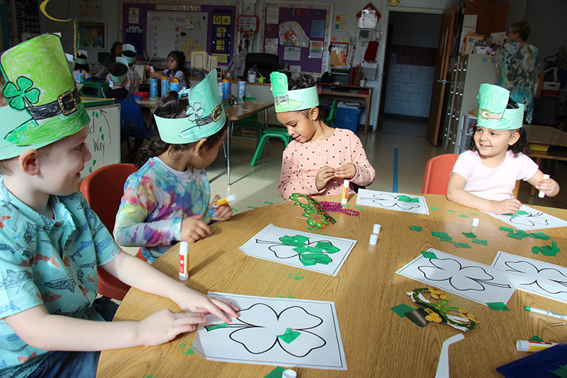 Four kids are sitting around a large desk, all with green headbands on, working on pasting green pieces of paper onto an outline of a four-leaf clover.
