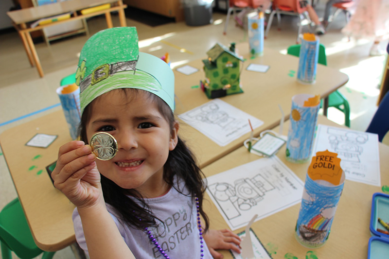 A preK girl with a green headband on holds up a large gold coin and smiles.