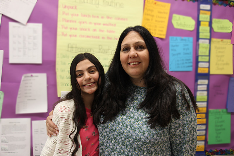 A woman on the right with long dark hair smiles. She has her arm around a young girl also with long dark hair. She is smiling too.