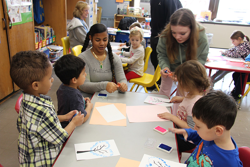 Two middle school-age girls sit at a table with pre-k kids helping them do a project.