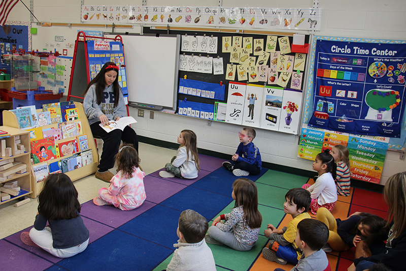A girl with long dark hair sits in front of a class of pre-K kids and reads to them.