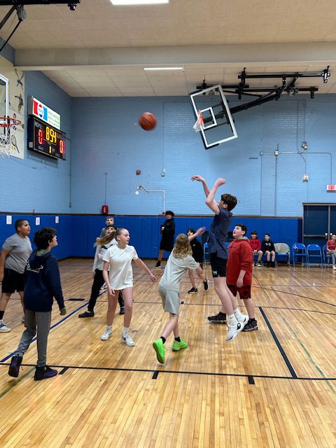 A group of middle school kids play basketball. A ball is shot toward the basket.