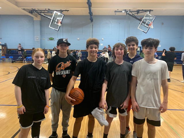 A group of six middle school kids smile and stand together. On kid is holding a basketball.