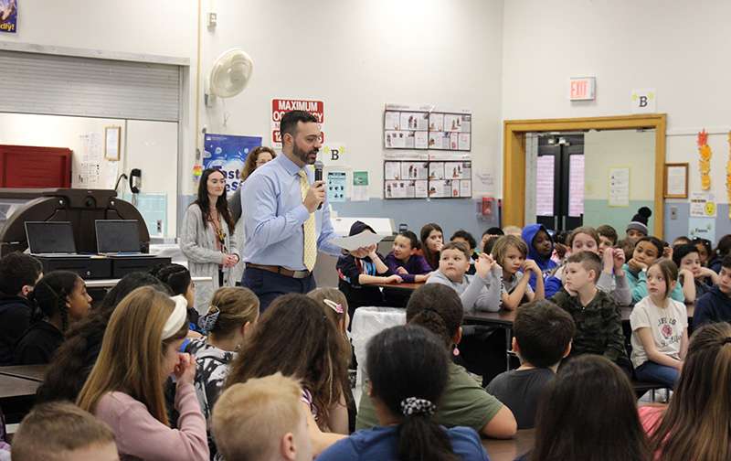 A man in a light blue shirt and yellow tie stands amid a large group of third and fourth grade students. He is holding a microphone talking to them.