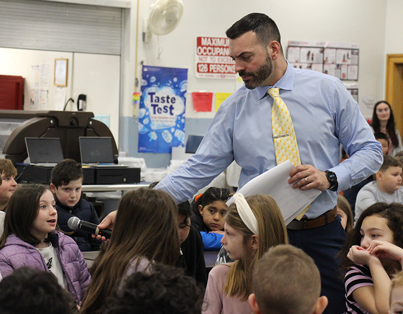 A man with dark hair a a dark beard wearing a blue shirt and yellow tie holds out a microphone to a young elementary student who is speaking to a large group.