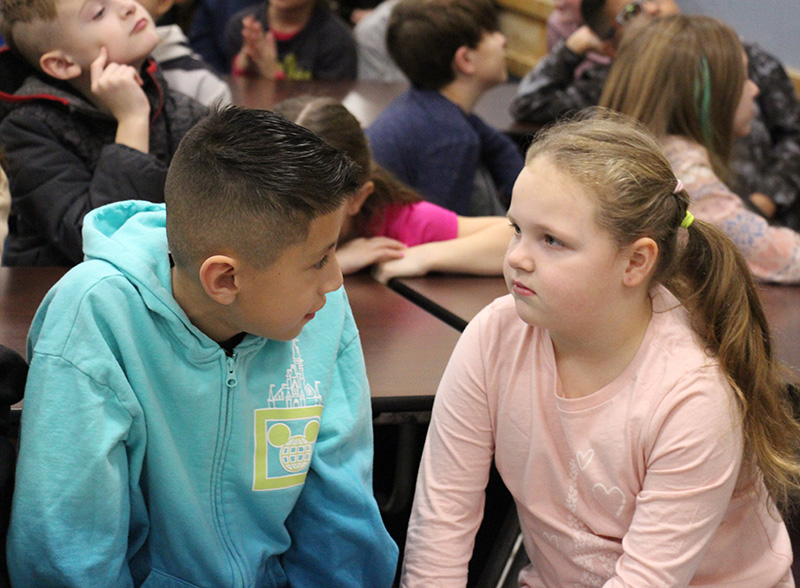 Two mid-elementary age students, a boy on the left with short dark hair wearing a blue hoodie, and a girl on the right with her light hair pulled back in a ponytail wearing a pink shirt, face each other and talk.