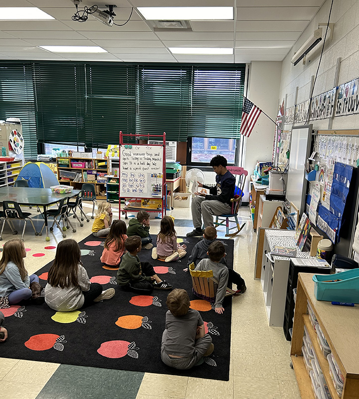 A high school student sits on a rocking chair in front of a group of kindergarten kids who are seated on a rug in front of him.