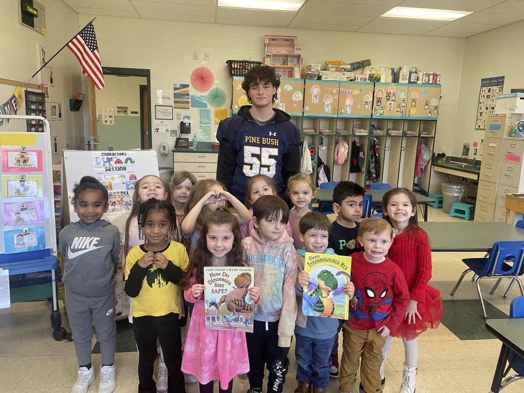 A high school football player stands with a group of prek students. They're all smiling and two kids are holding up books the player just read.