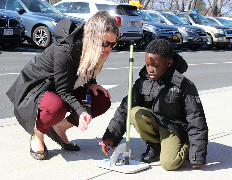 A woman with long blonde hair squats down to help a student put a straw rocket on a launcher. 