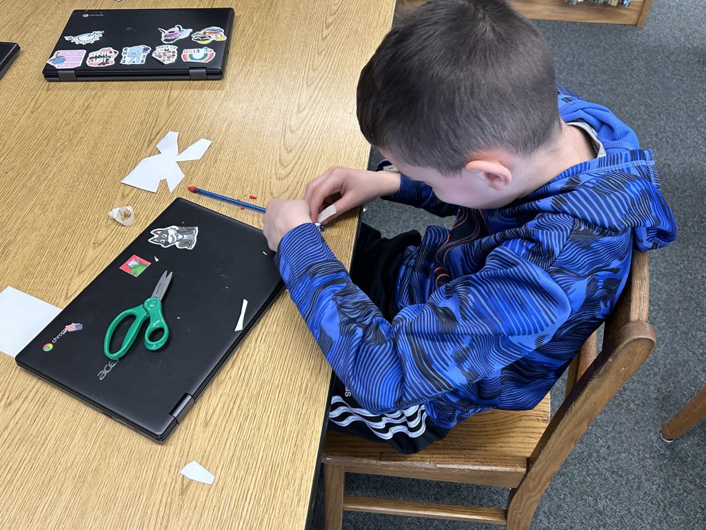 An elementary-age boy sits at a table making a straw rocket.