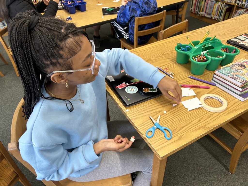 An elementary age girl sits at a table making a straw rocket.