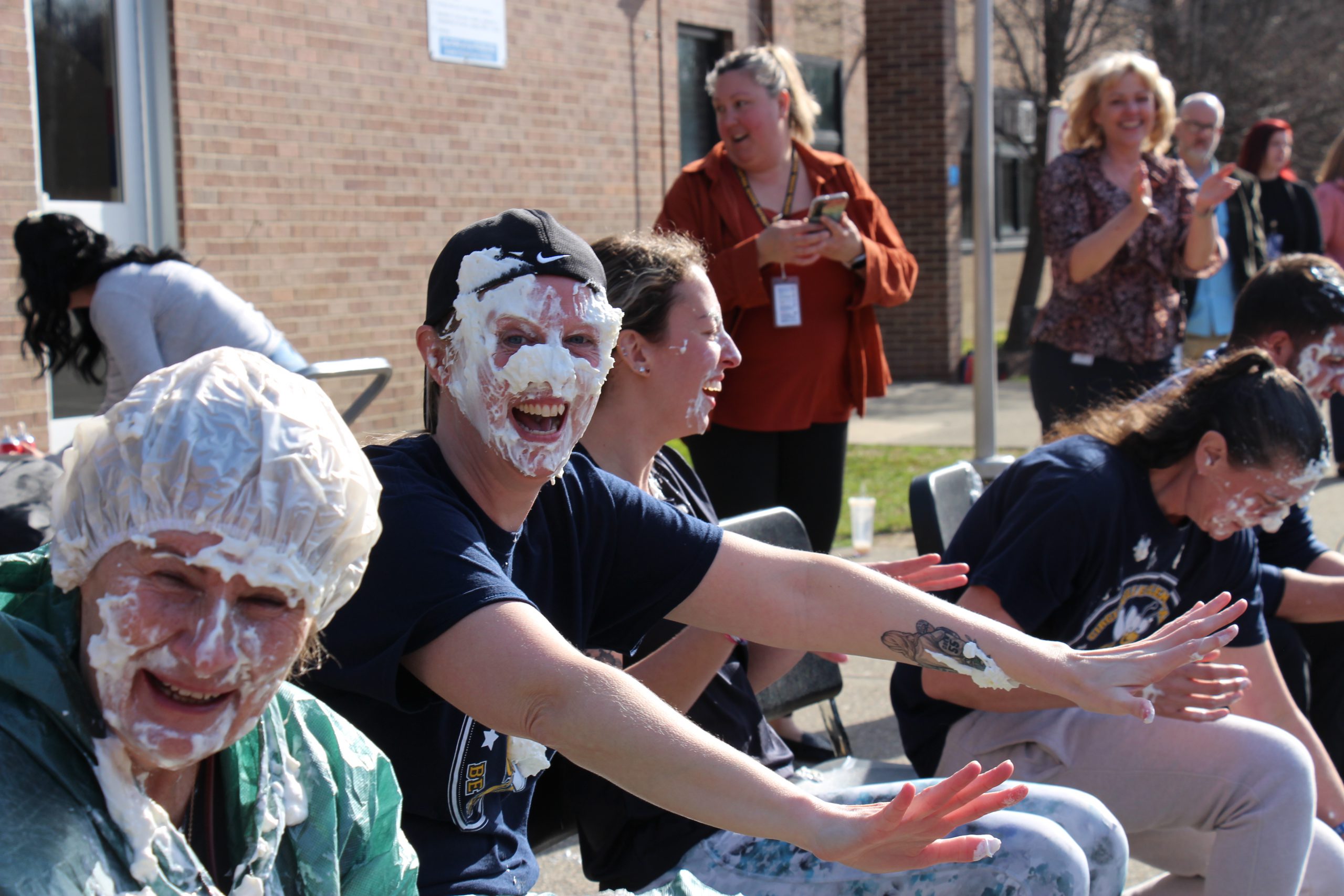 A woman with a backward baseball cap smiles with white whipped cream all over her face.