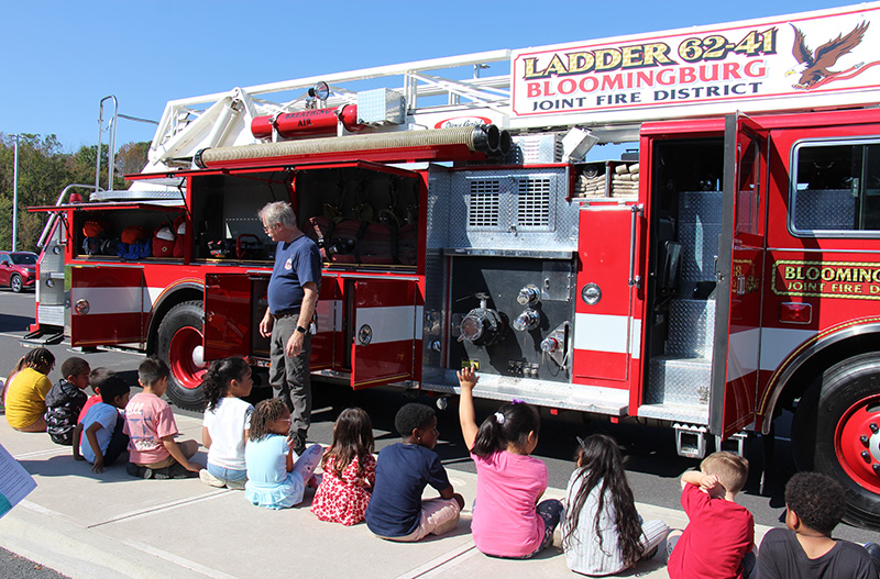 A man stands in front of a group of elementary students with a red fire truck behind him.