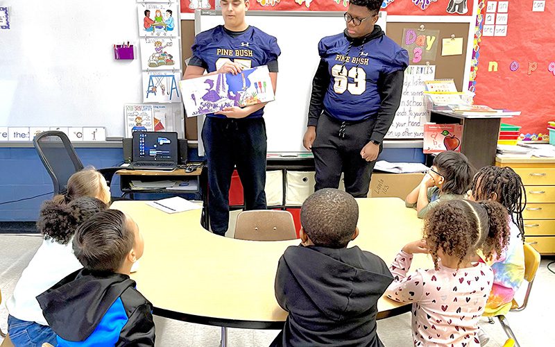 Two high school football players wearing blue jerseys read to a group of elementary students seated at a desk in front of them.