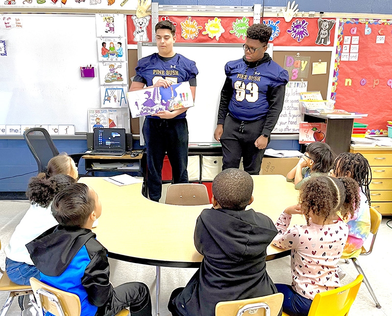 Two high school football players wearing blue jerseys read to a group of elementary students seated at a desk in front of them.