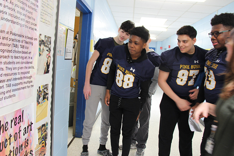 A group of five high school football players wearing their jerseys stand in a hallway and look at photos on a wall from when they were in elementary school.