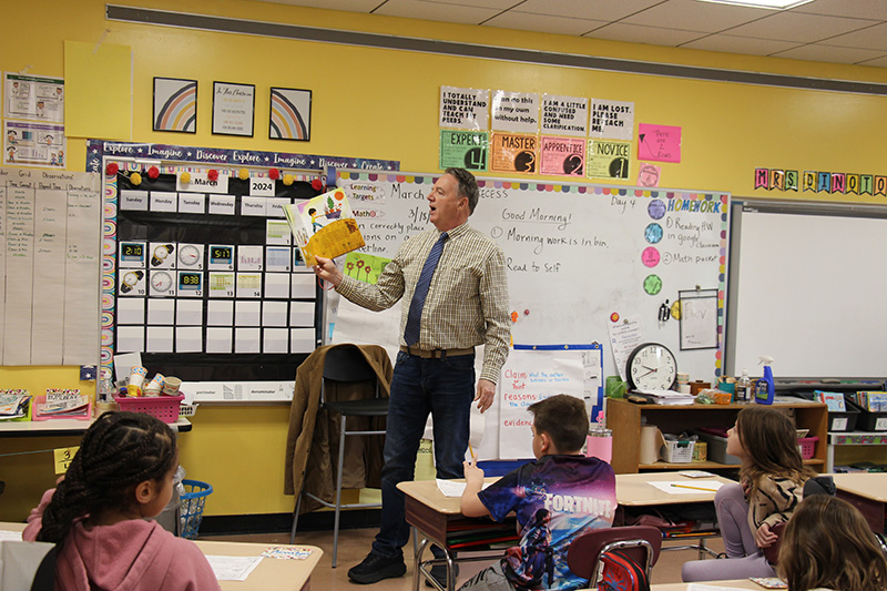 A man wearing a shirt and tie stands in front of a classroom reading a book.