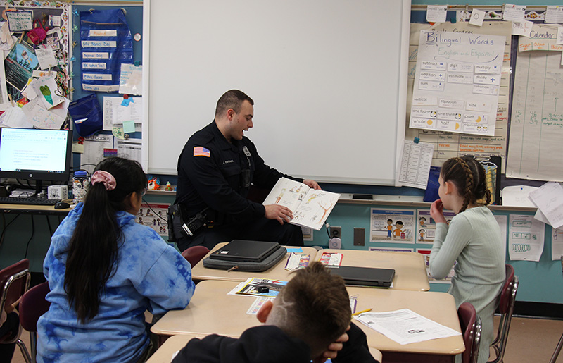 A male police officer sits in front of a class reading a book.