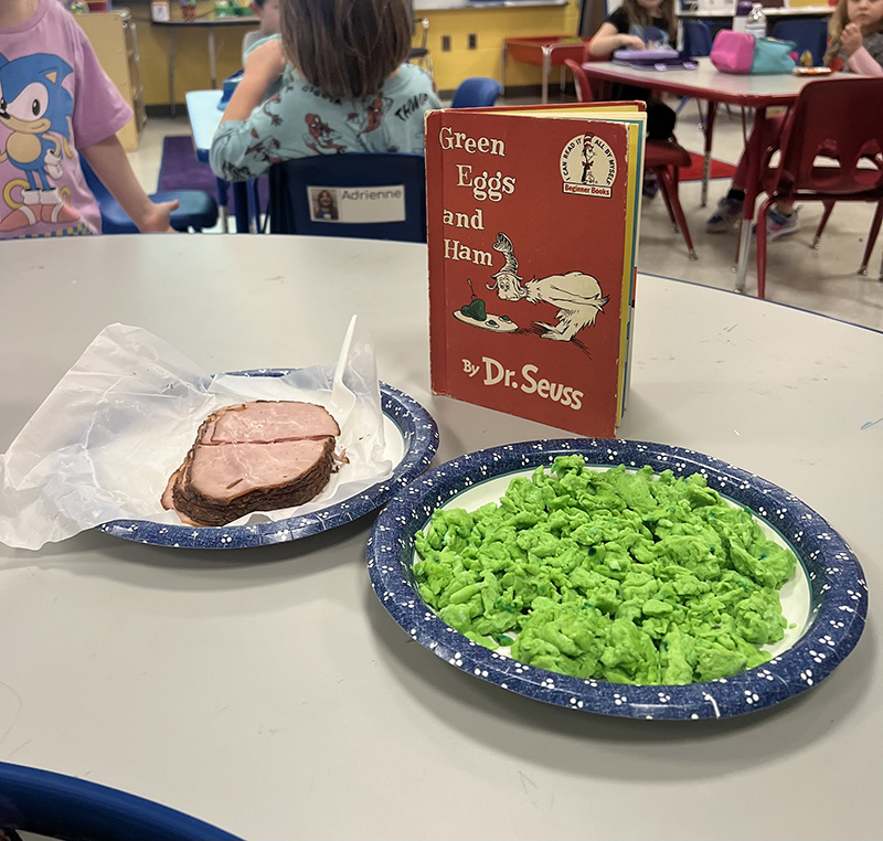 A book stands on a table and in front of it is a plate with green scrambled eggs and another with cooked ham.