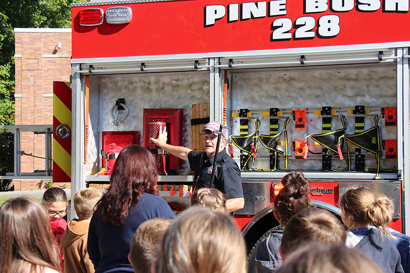 A man stands in front of a red fire truck that says Pine Bush 228. Several students are in front of him listening.