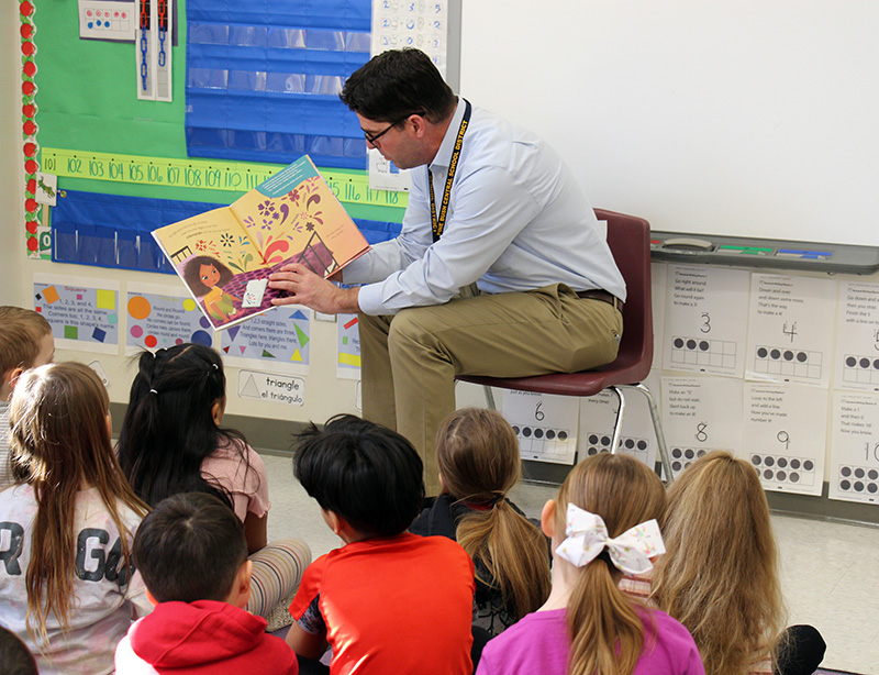 A man wearing a blue shirt and khaki pants sits and reads a book to a class of pre-K students.