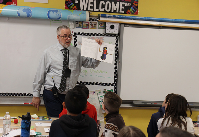 A man with glasses, wearing a shirt and tie holds up a book as he reads it to his students.