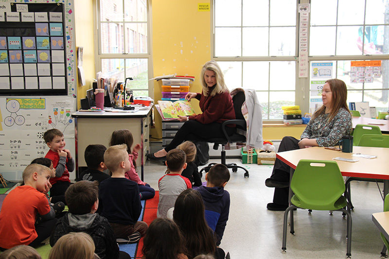 A woman sits in front of a class and reads a book to them.