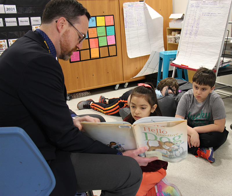 A man with glasses and short dark hair sits and reads to two children. He holds the book down so a girl can look at it.