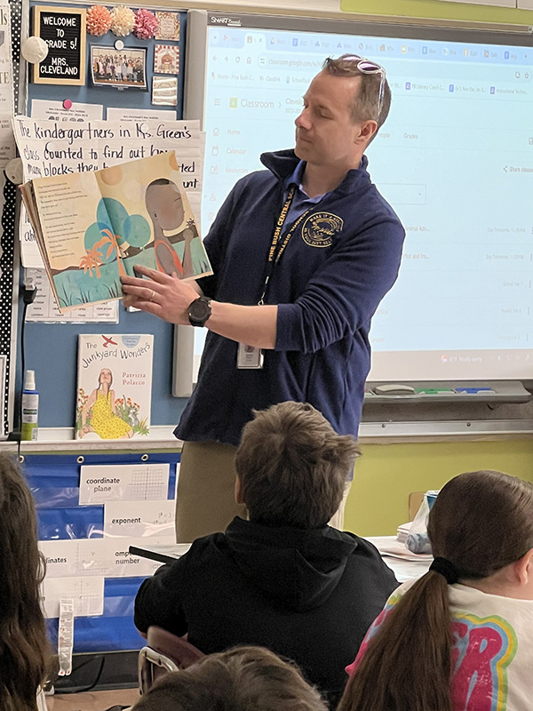 A man in a blue jacket holds up a book as he reads it to a class.