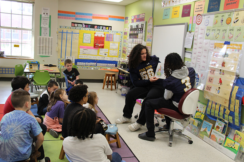 Two high school football players wearing blue jerseys read to a group of elementary students who are sitting on a rug in front of them.