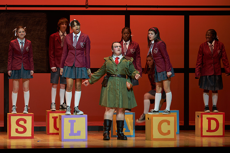 On a stage, actors standing on large lettered blocks. They are wearing gray skirts and maroon blazers. In the center is a character wearing a green dress and red tie.