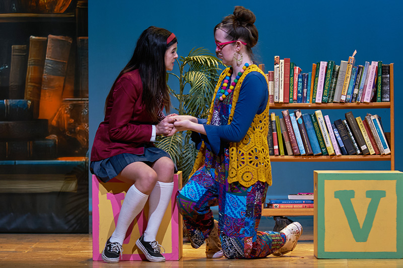 Two actors - a girl sits on a large block and another kneels down next to her. They are holding each other's hands.