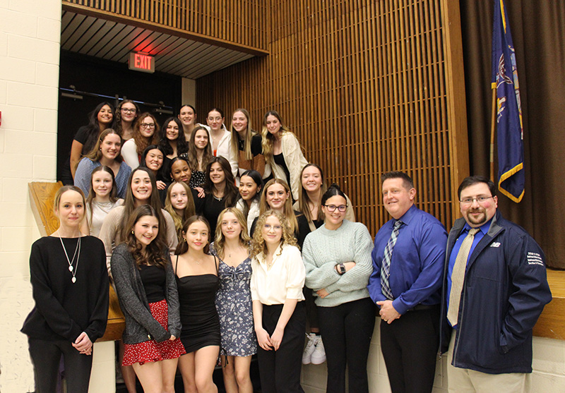 A large group of high school girls stand together, going up a flight of stairs. With them are three adults. All are smiling.