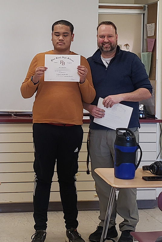 A young man smiles and holds a certificate as he stands next to a man with dark hair and a mustache.