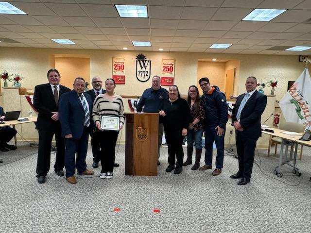 A group of nine adults and one young woman standing around a podium at a municipal meeting.