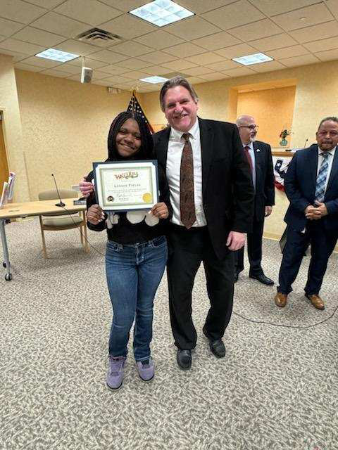 A high school girl smiles as she holds a certificate. She is standing with a man in a suit and tie who is smiling too. 