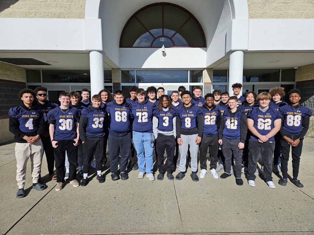 A group of 26 high school football players stand outside of building on a sunny day. they are all wearing blue football jerseys.