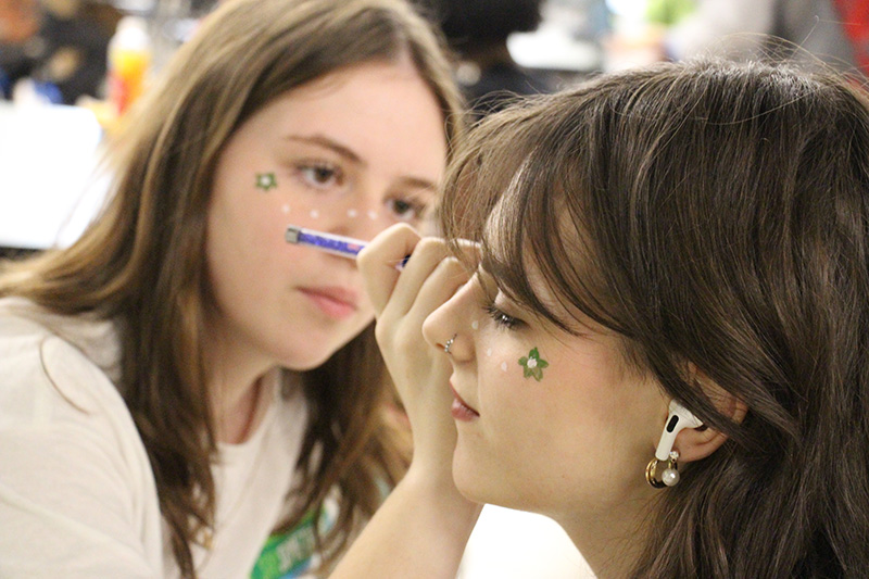 A high school girl sits while getting her face painted by another high school girl. 