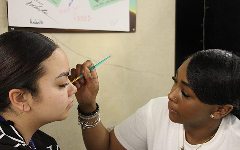 A high school girl paints the face of another high school student.