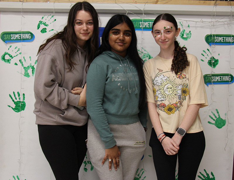 Three high school girls stand in front of a green and white banner that says Say Something.