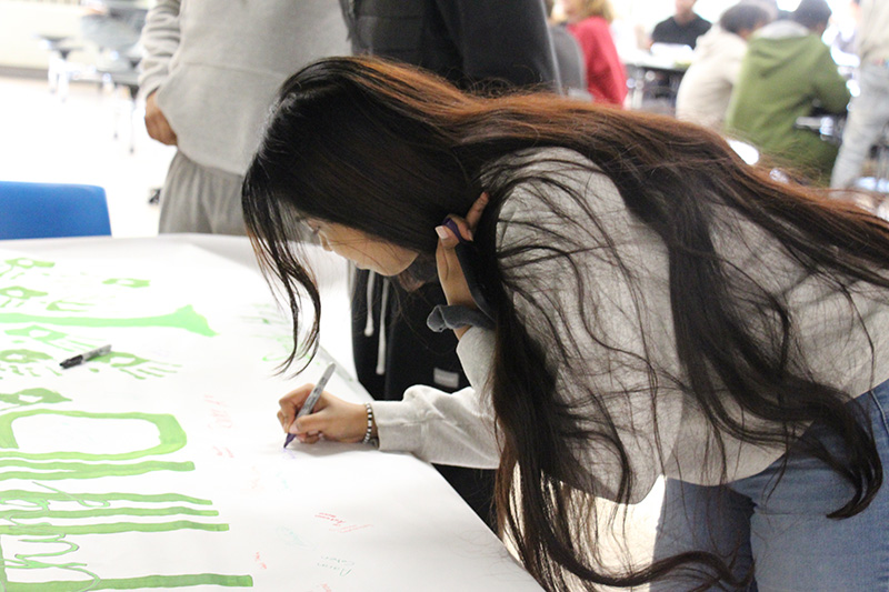 A high school girl with very long dark hair holds her hair back while she signs a large poster on a table.