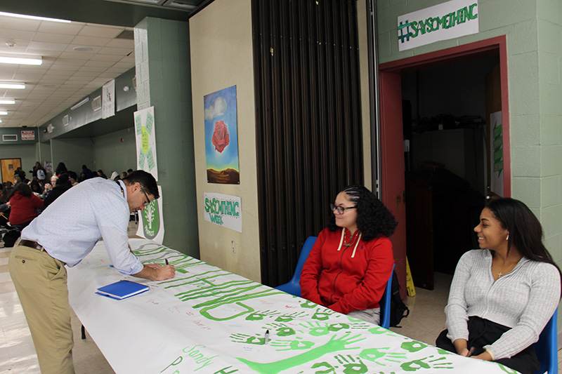 A man leans over a table and signs a large green and white sign as two high school girls sit and watch him.