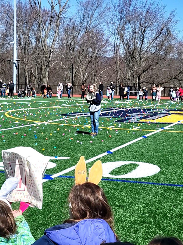 On a green football field, a woman holds a megaphone to talk to the many people who are lined up to find eggs.