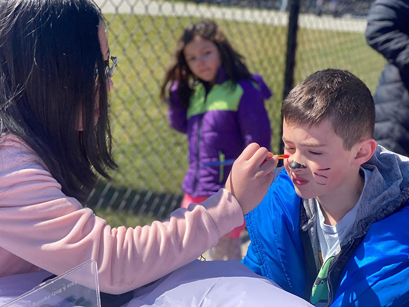 A boy sits and gets his face painted by a high school girl.