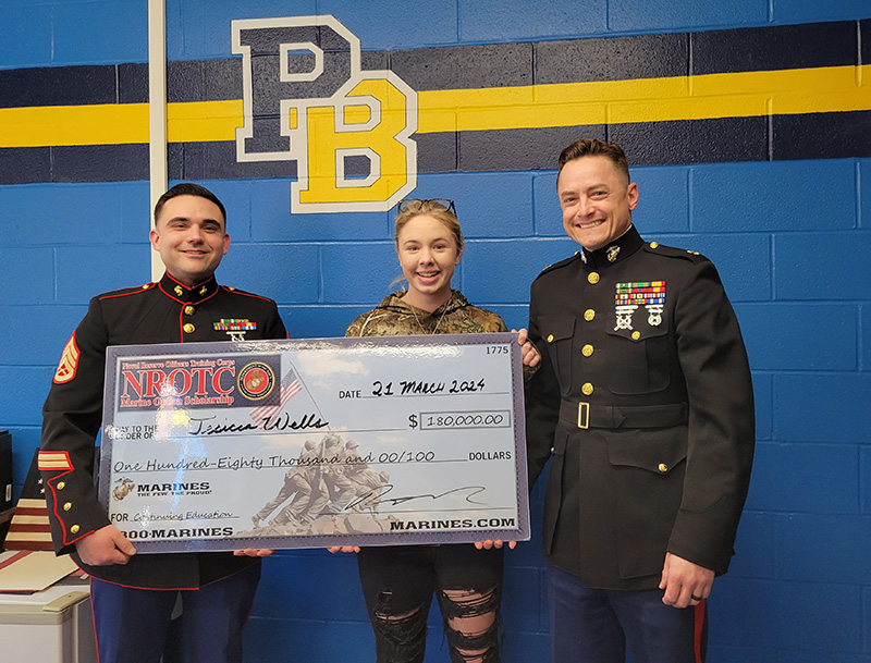 A high school girl stands in the center of two men in marine uniforms. They are holding an oversized check for $180,000.