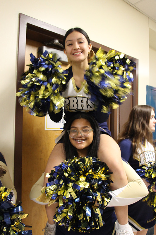Two high school cheerleaders, one sitting on the shoulders of the other. They have blue and gold pom poms.