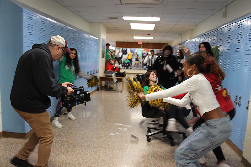 A man on the left holding a video camera while kids on the right wave to him.