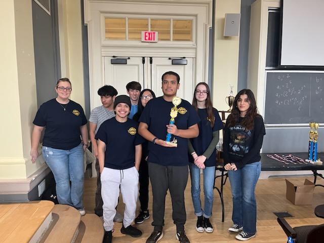 A woman and seven high school students stand together smiling. The young man in front holds a trophy.