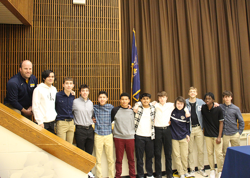 A group of 11 high school boys stand together in front of a stage and up a short set of stairs, with a man on the left.