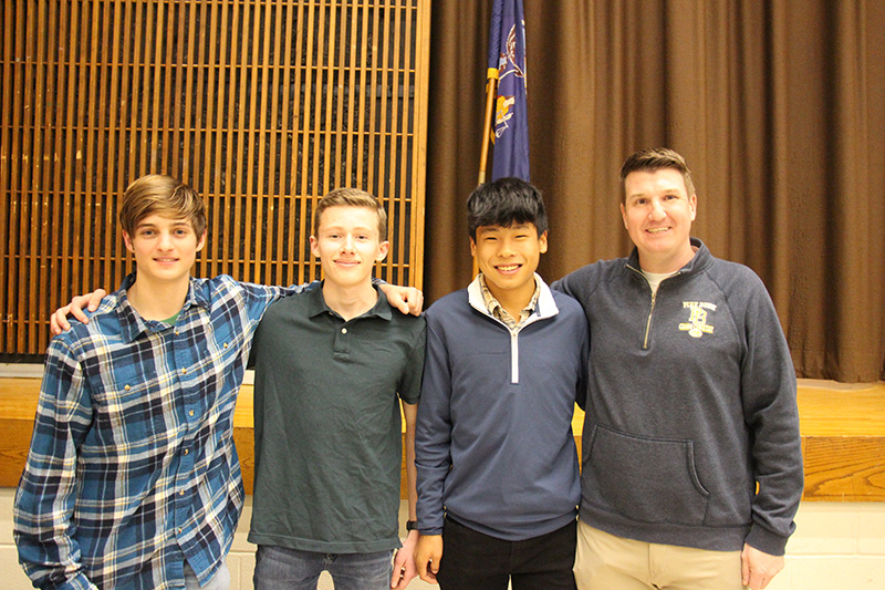 A man stands on the right wearing a blue sweatshirt and three young men stand next to him. They are all arm-in-arm.